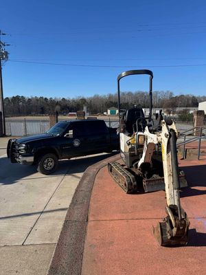 A black truck is parked next to a small excavator on a sidewalk.