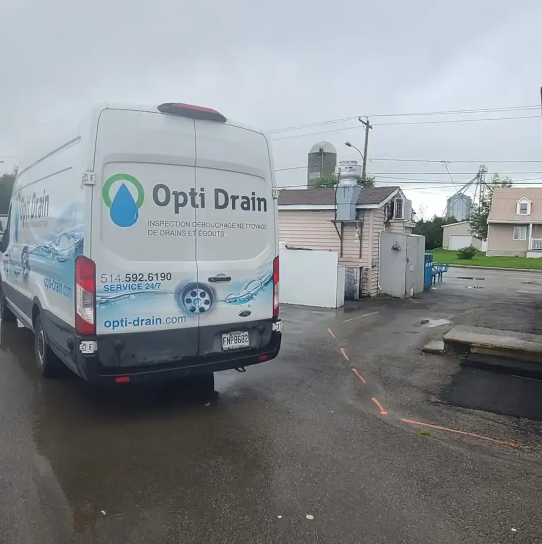 Une camionnette blanche avec le mot opti drain à l'arrière est garée sur un parking.