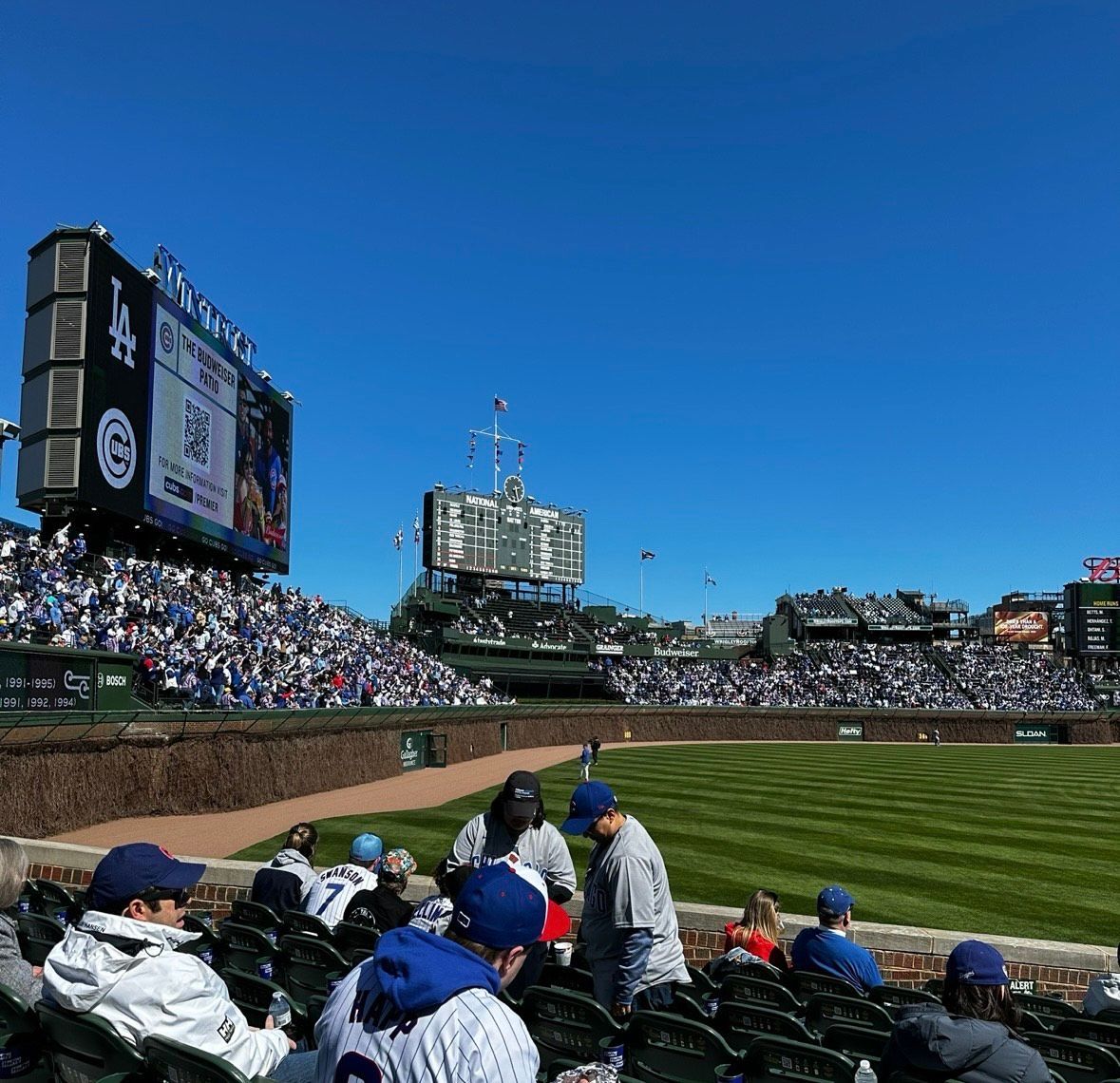 Baseball game at Wrigley Field: Fans in blue hats watch the game on a sunny day. Large scoreboard visible.