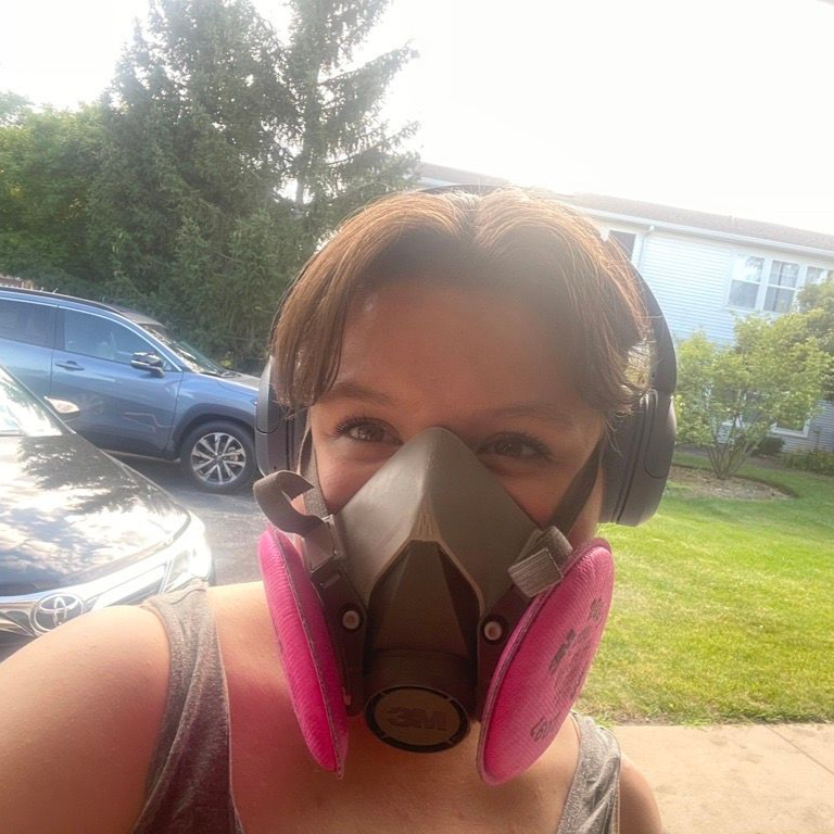 Woman wearing a respirator and ear protection outdoors, smiling in front of a house and cars.