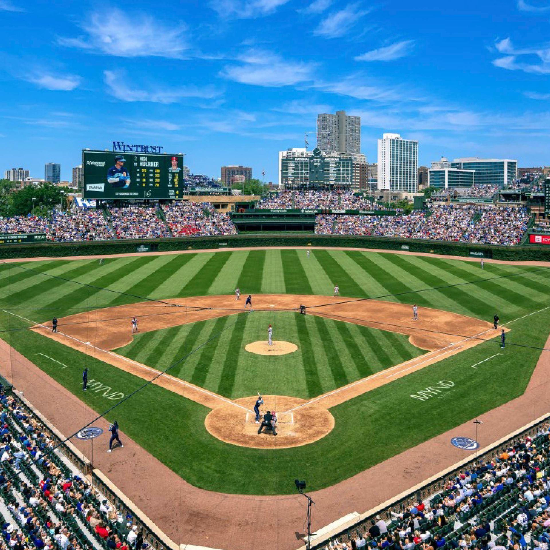 Baseball game at Wrigley Field: Fans in blue hats watch the game on a sunny day. Large scoreboard visible.