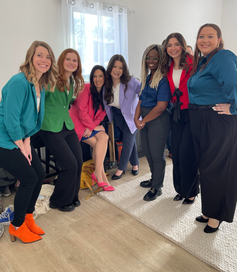 Eight people smiling and posing in a room. They wear colorful blazers with various outfits. White walls, window, and rug visible.