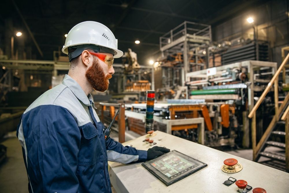 A man is sitting at a control panel in a factory looking at a tablet.