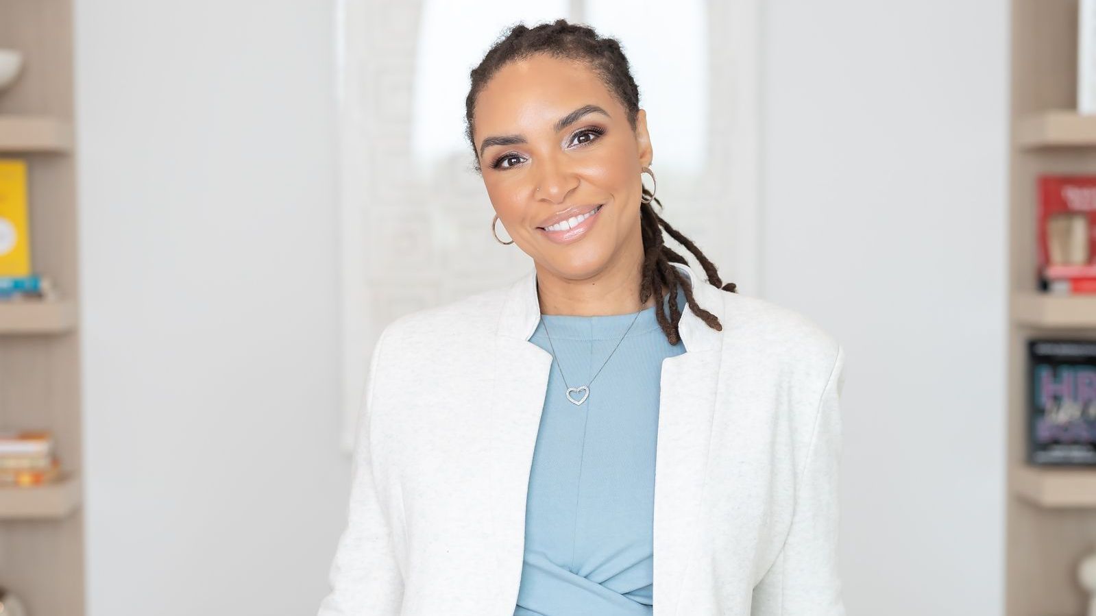 Woman with locs smiles, wearing a white blazer and blue top, in a well-lit room with shelves.