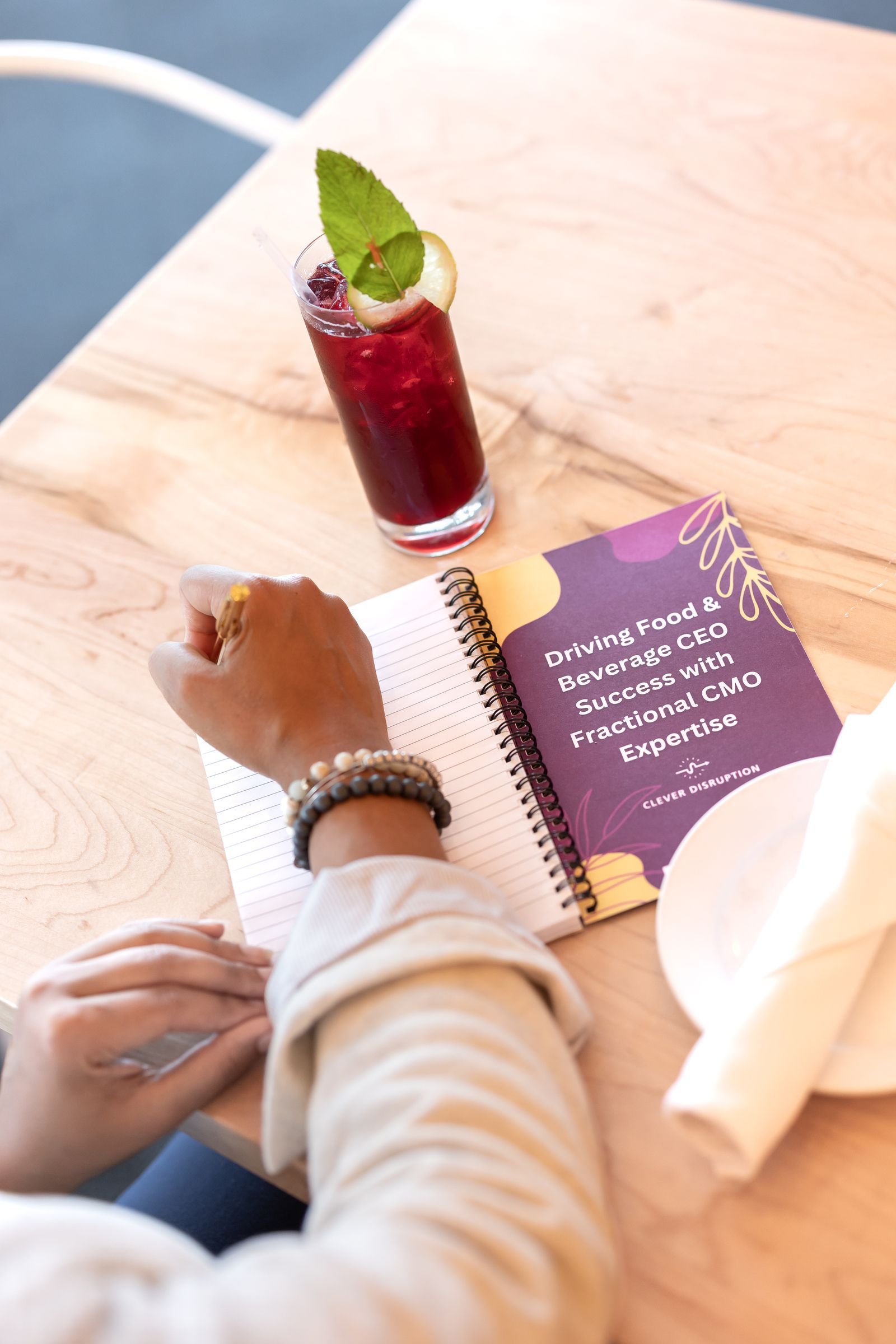 A person writing in a notebook at a table with a cocktail. The notebook is open next to a drink and a rolled napkin.