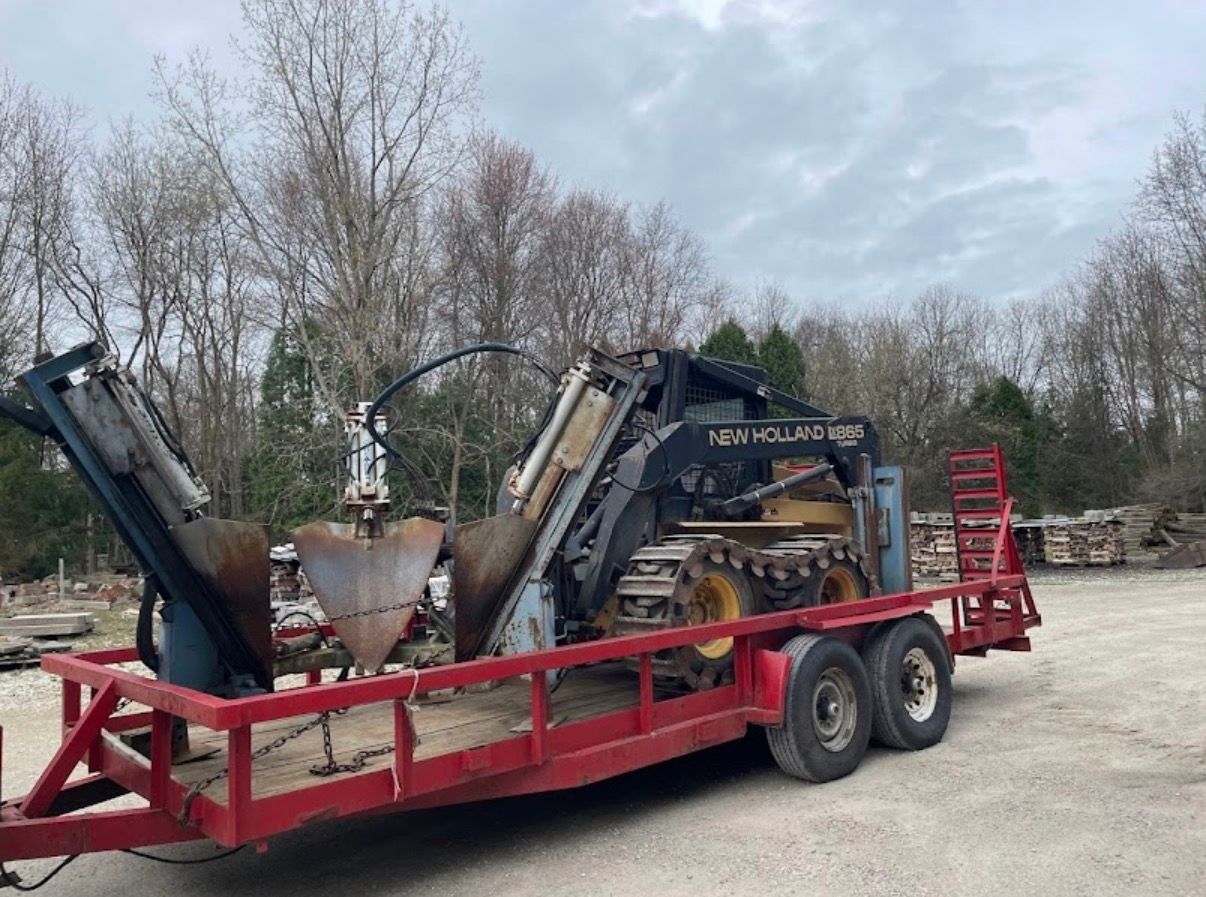 A tracked loader with a tree spade attachment mounted on a red flatbed trailer outdoors against a backdrop of trees.