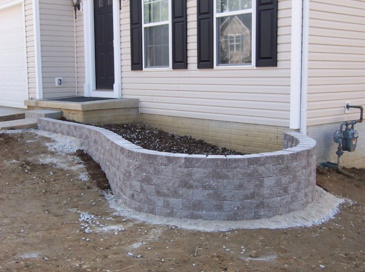 A newly built curved gray stone retaining wall in front of a house, filled with dark soil and bordering a porch.