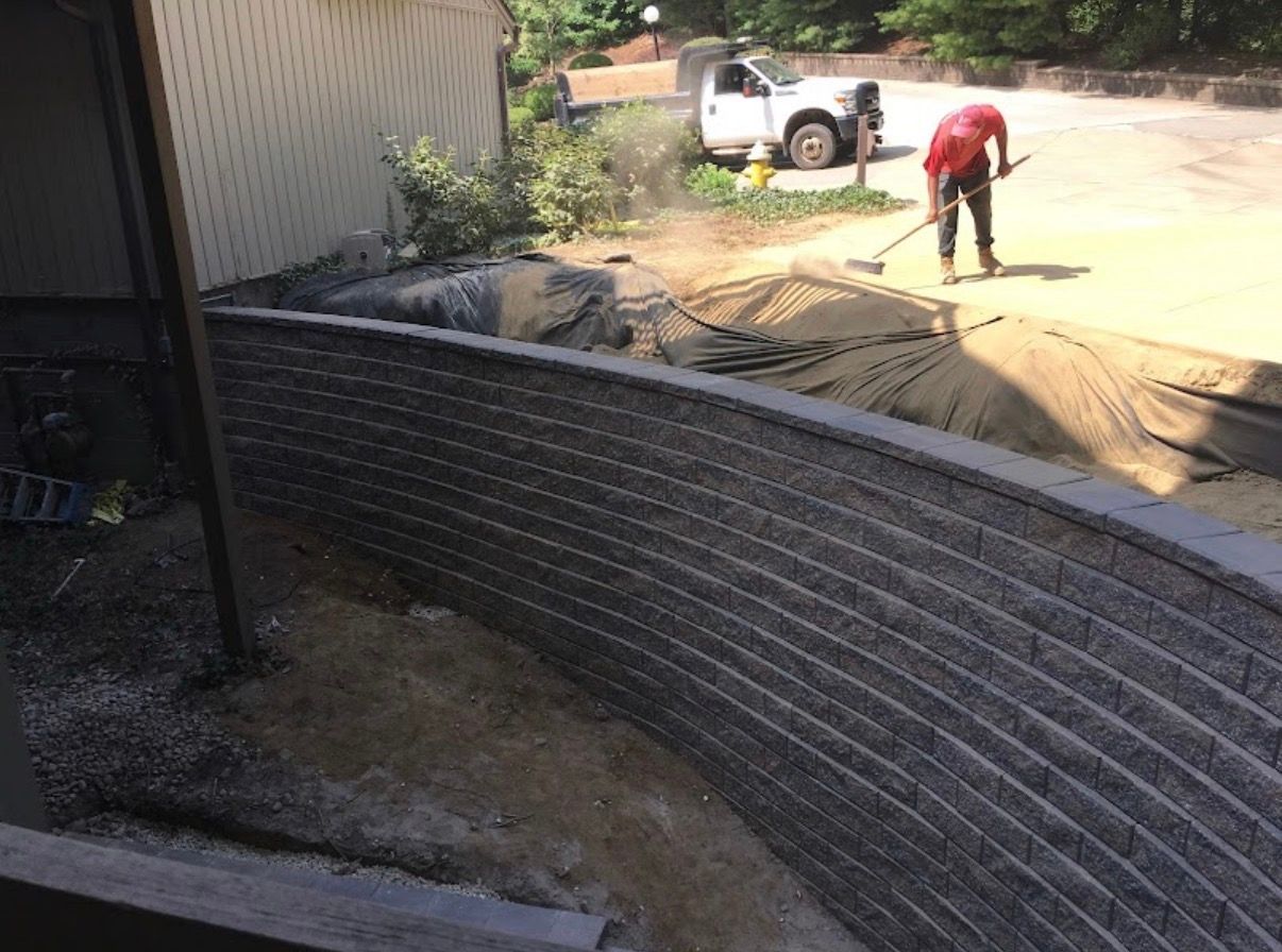 A worker spreads gravel with a rake near a newly constructed curved stone retaining wall on a sunny day.