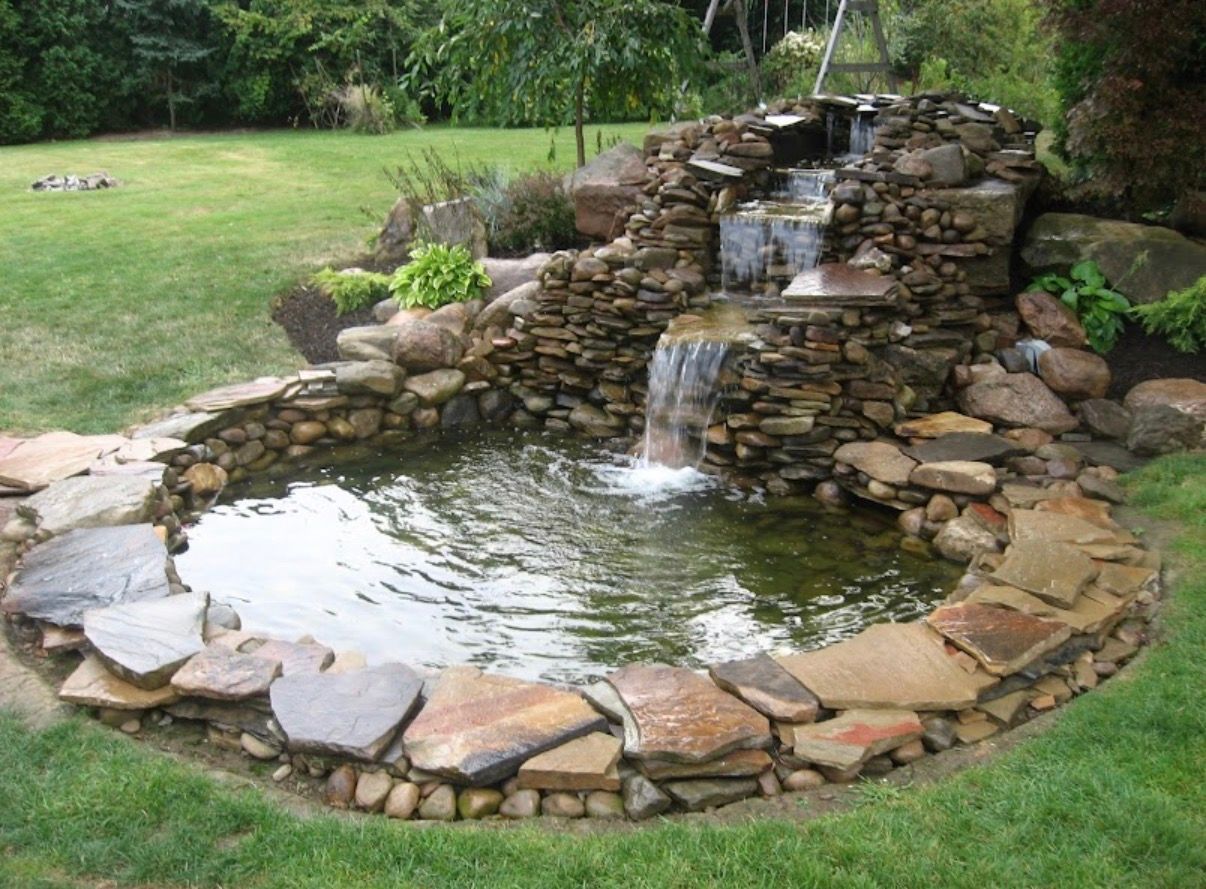 A backyard stone pond with a multi-tiered rock waterfall, surrounded by grass and landscaping stones.