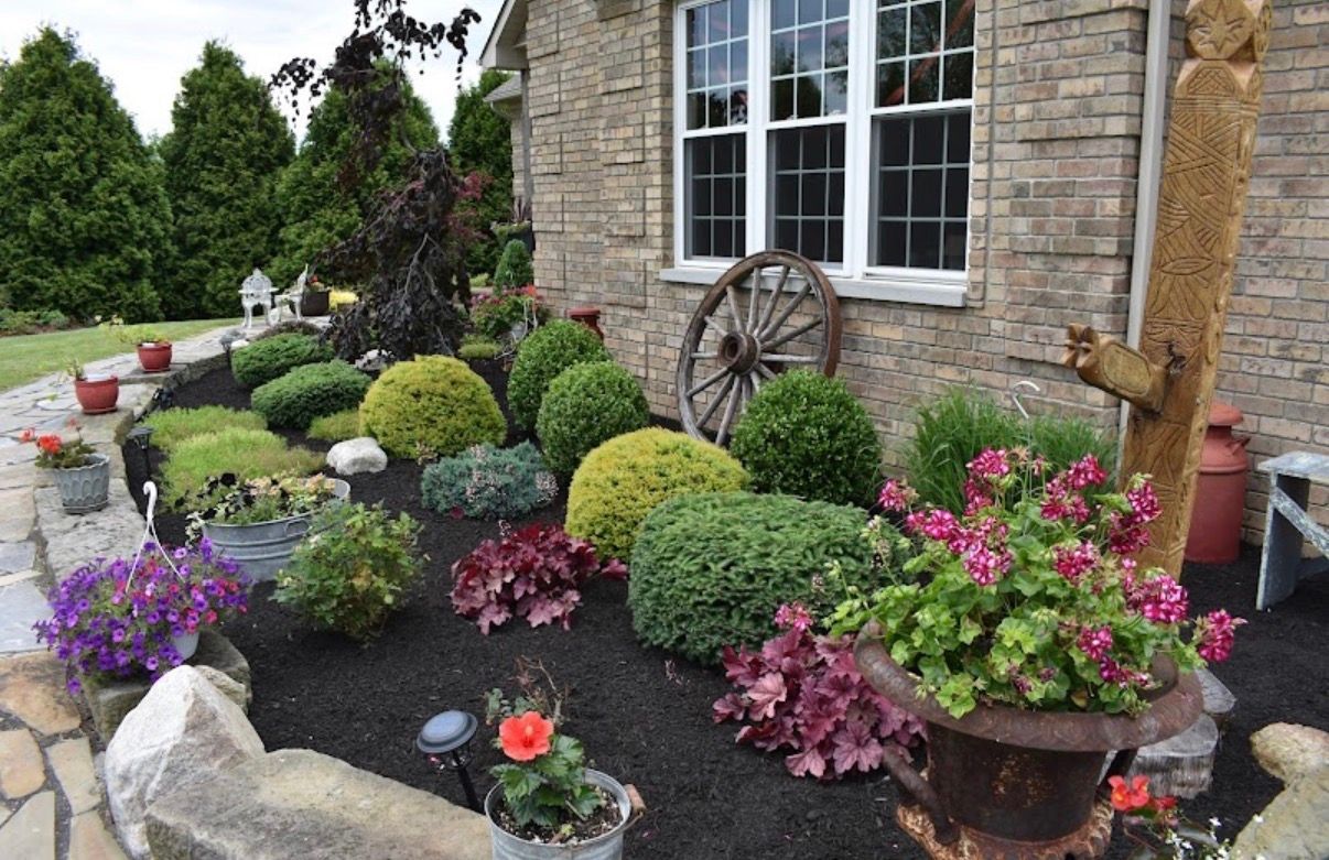 A landscaped garden bed in front of a stone house featuring shrubs, colorful flowers in pots, and a vintage wagon wheel.