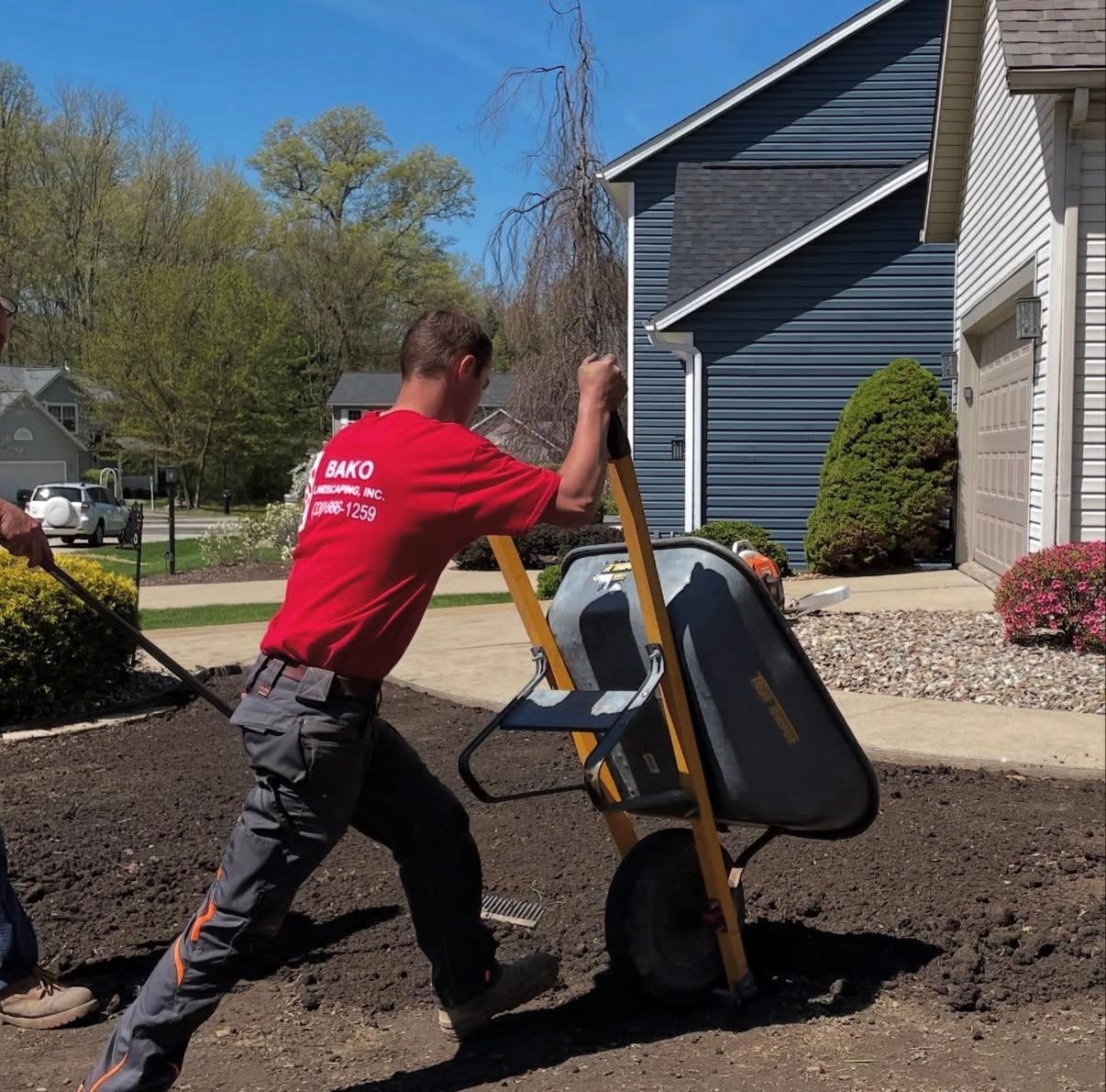 A person of Bako Landscaping wearing a red t-shirt and grey work pants pushes a wheelbarrow
