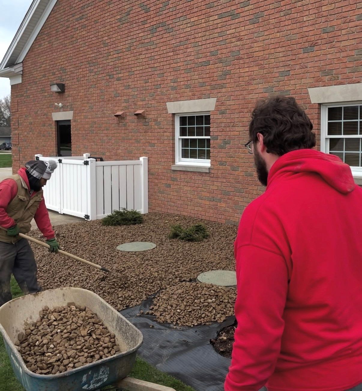 Two people of Bako Landscaping work on landscaping, spreading gravel over a weed barrier beside a brick building.