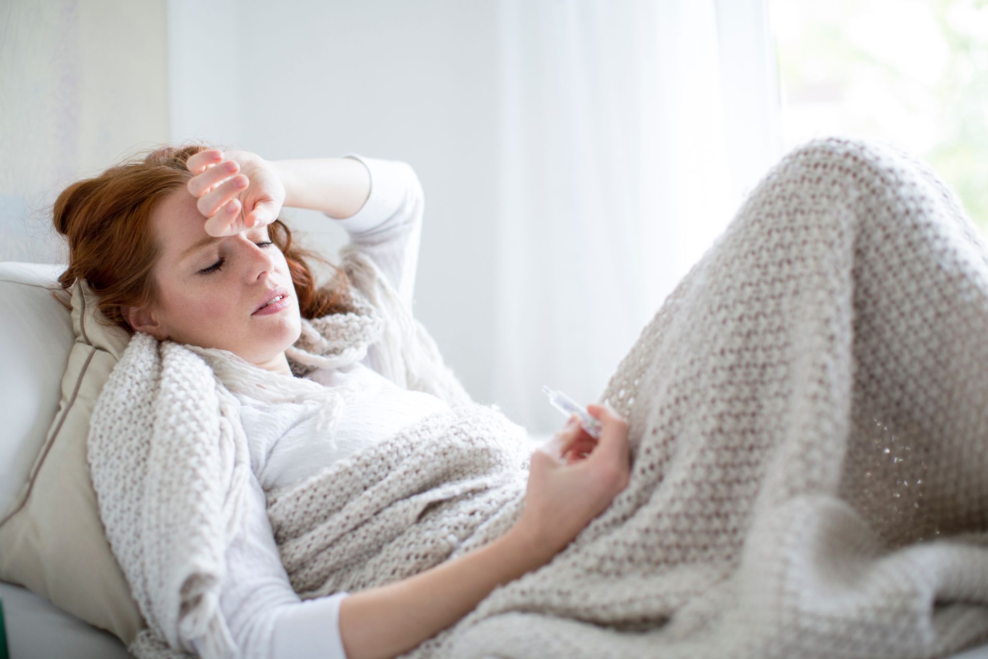 Eine Person liegt im Bett unter einer Decke, eine Hand auf der Stirn, in der anderen hält sie ein Thermometer.