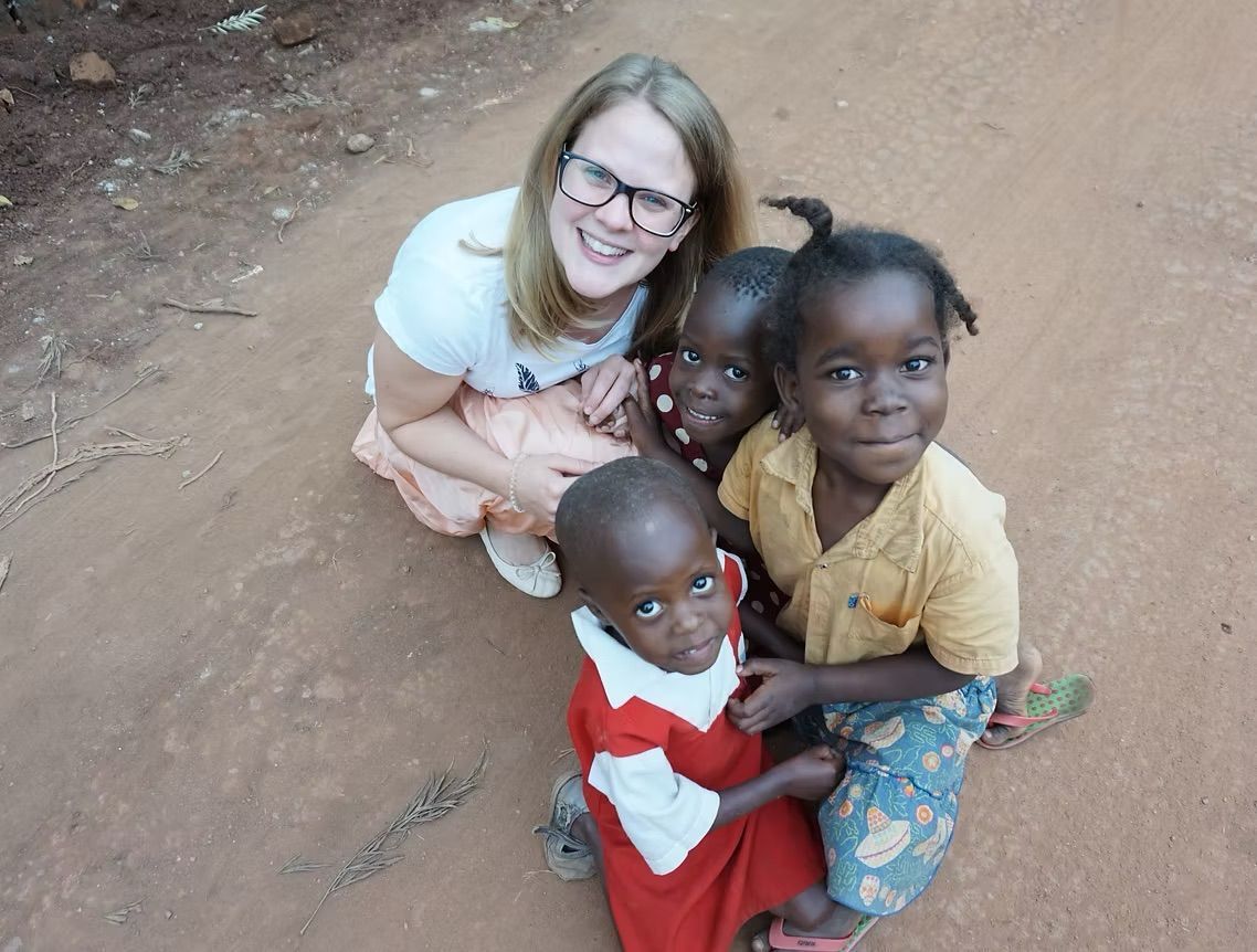 Eine lächelnde Person hockt auf einem Feldweg mit drei Kindern, die ebenfalls in die Kamera lächeln.