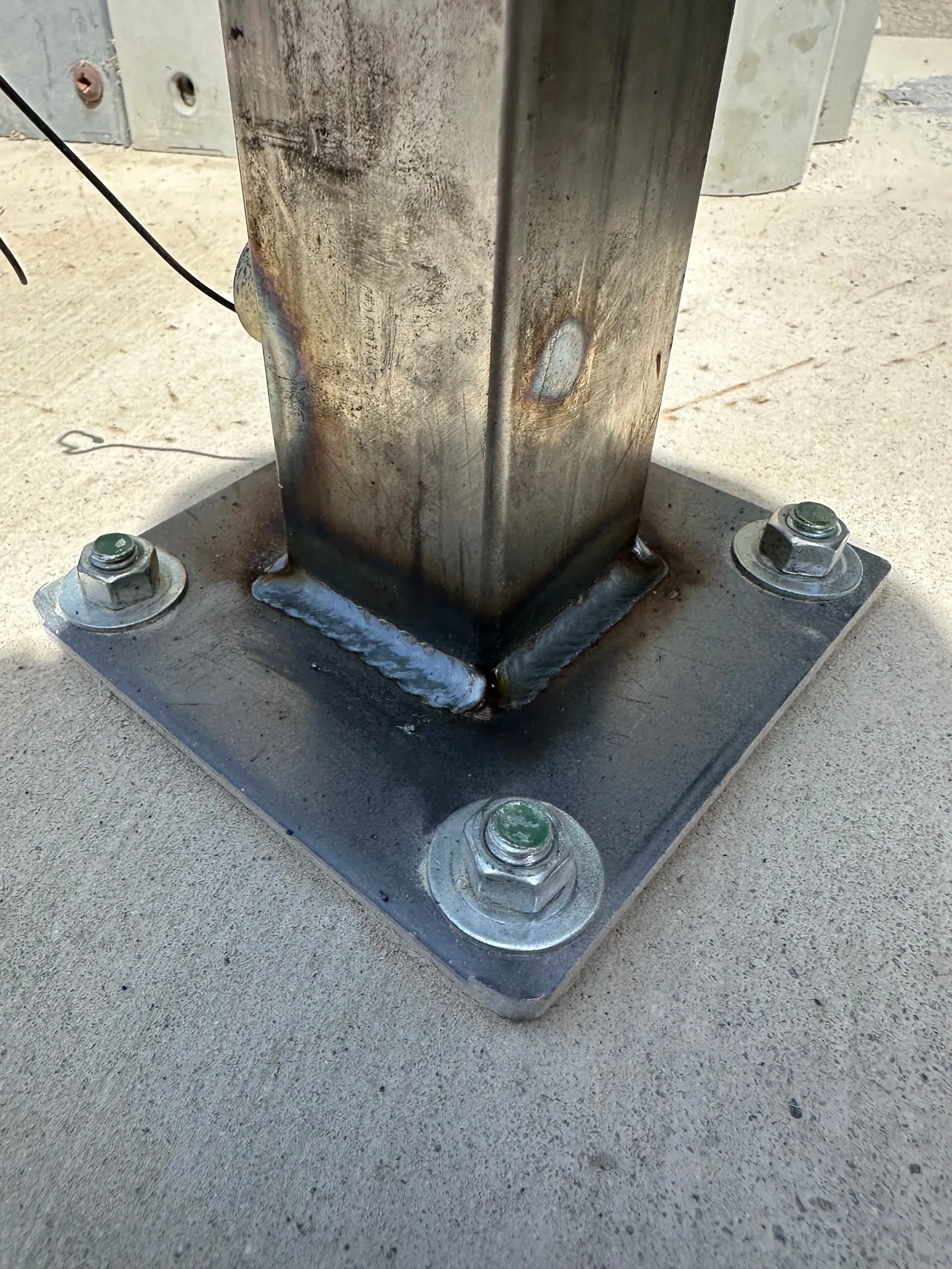 Steel post base plate bolted to concrete, with welded support at the bottom.