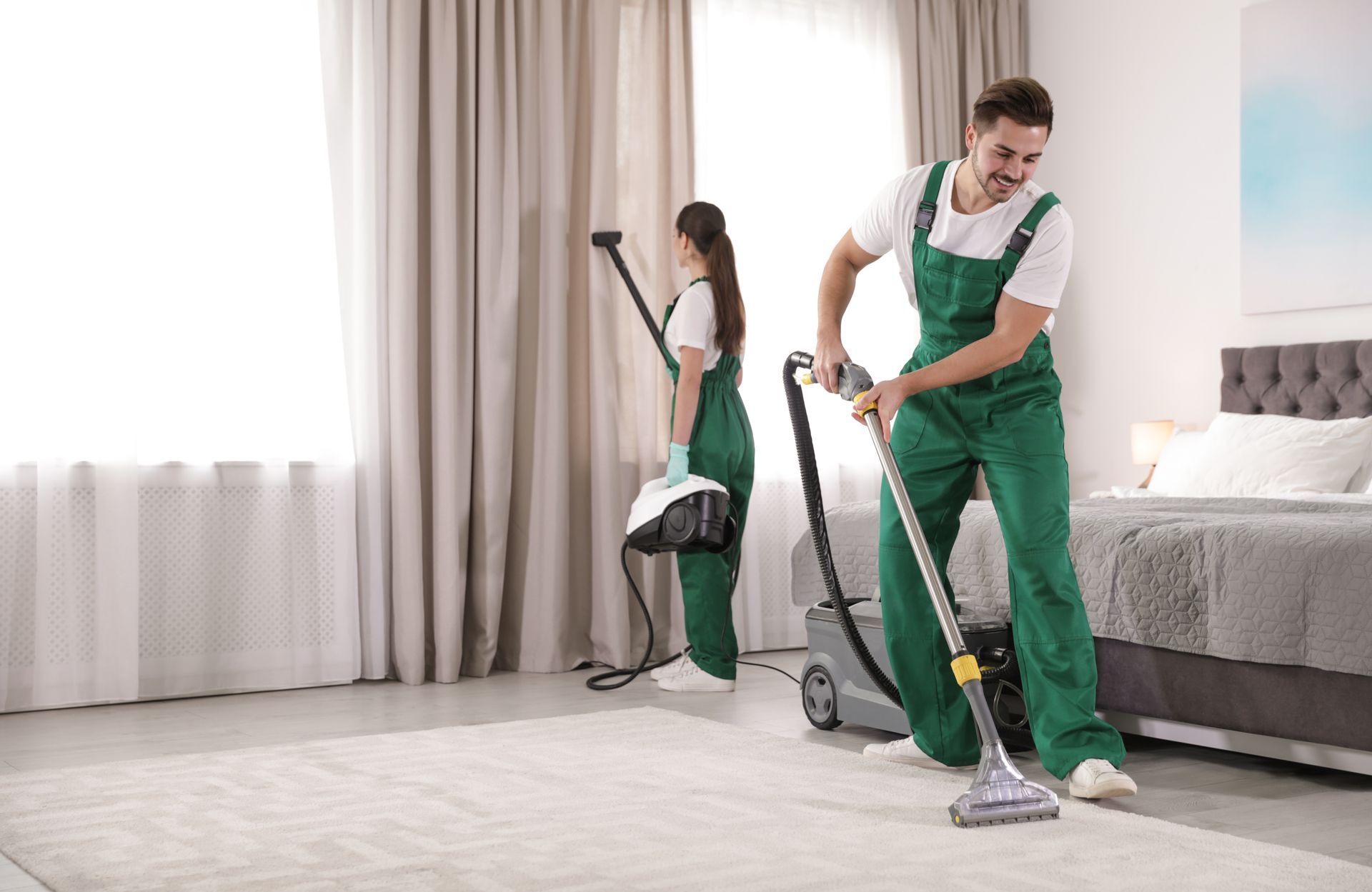 Two people in green overalls cleaning a bedroom carpet with vacuum equipment.