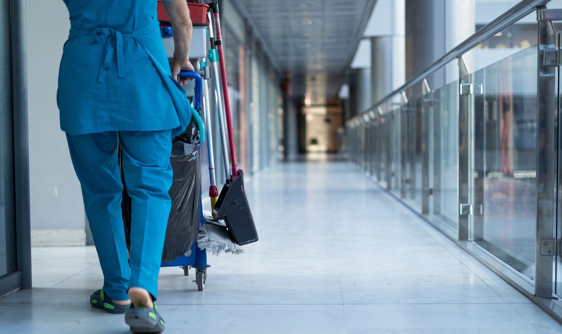A person in blue scrubs pushing a cleaning cart down a brightly lit hallway.