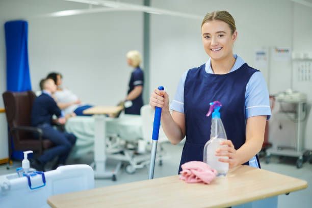 Hospital worker in blue uniform, smiling, holding cleaning supplies in a patient room.
