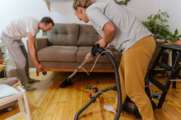 Two people vacuuming under a couch on a hardwood floor.