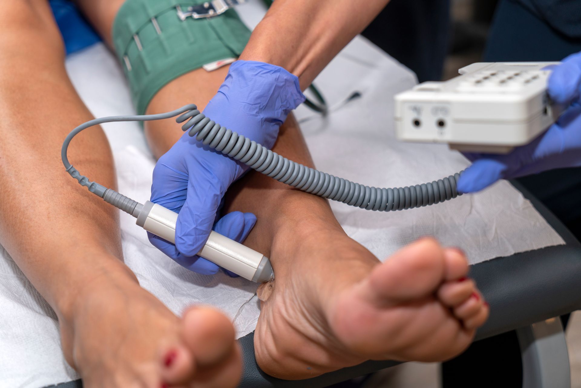 A healthcare provider in blue gloves uses a handheld Doppler ultrasound device to check the pulse on a patient’s ankle.