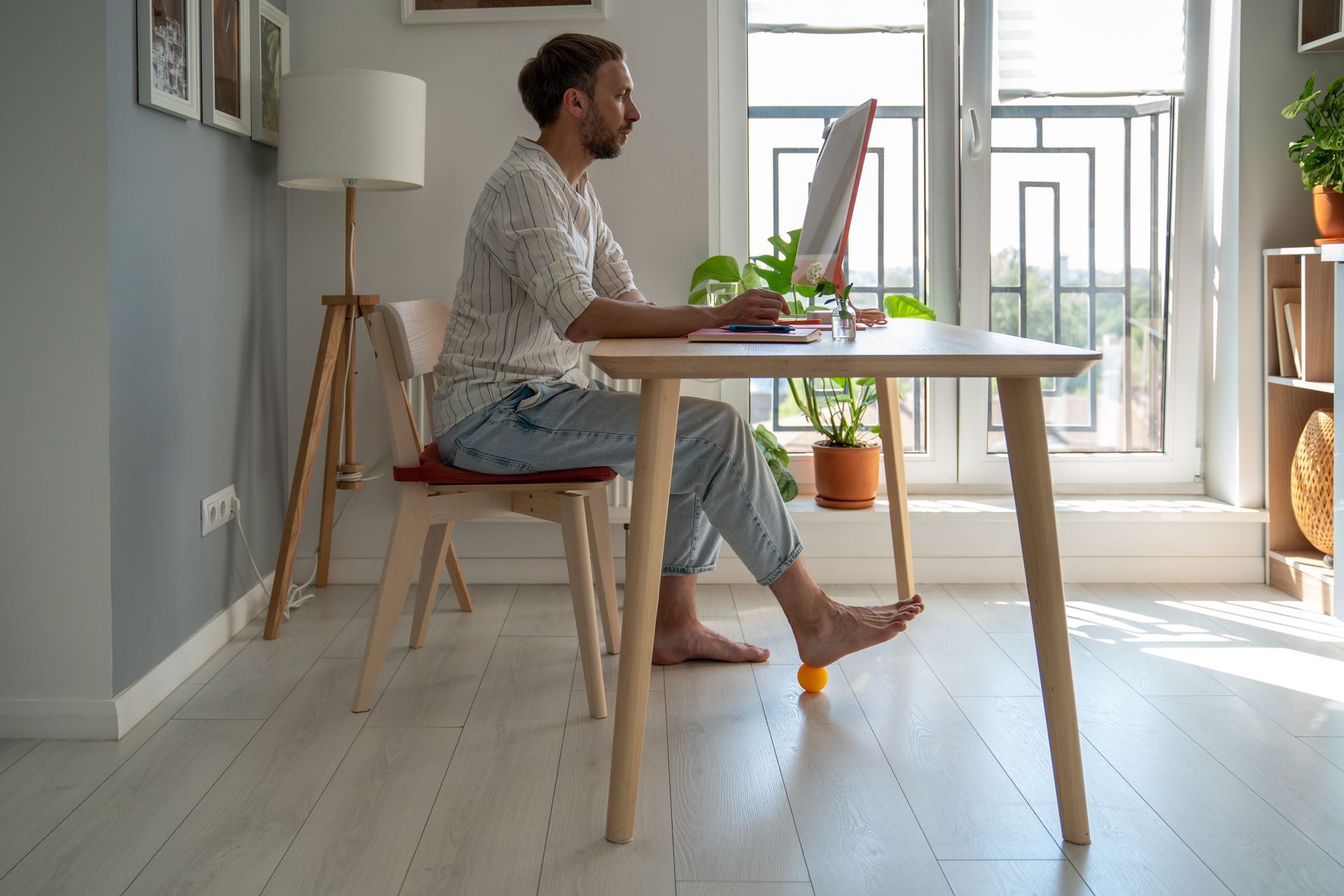 Person working on a laptop at a wooden table in a bright home office by a window.