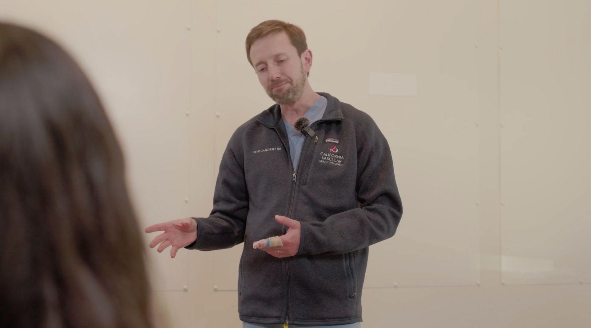 Man gesturing while speaking in a plain room, wearing a dark zip-up jacket