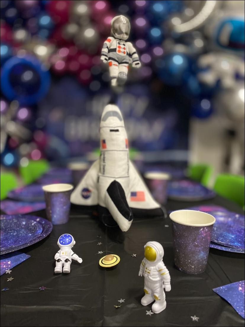 A table with plates , cups , napkins and a toy shuttle on it.
