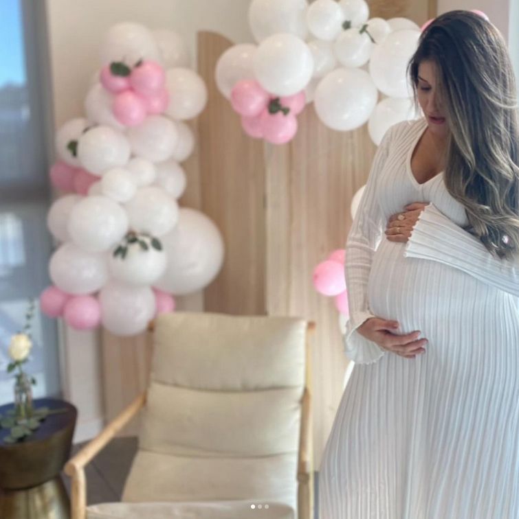 A pregnant woman in a white dress is standing in front of balloons.