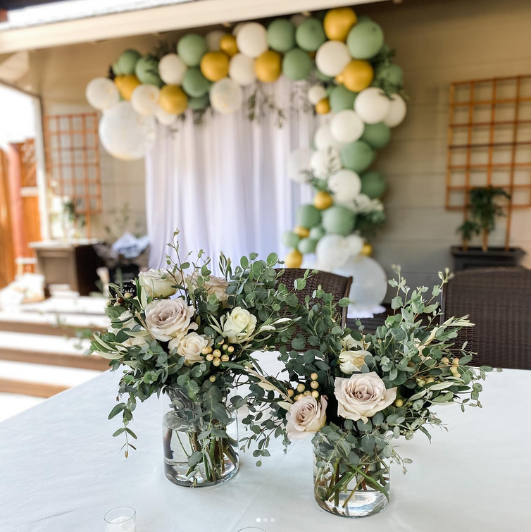 Two vases of flowers are on a table in front of a balloon arch.