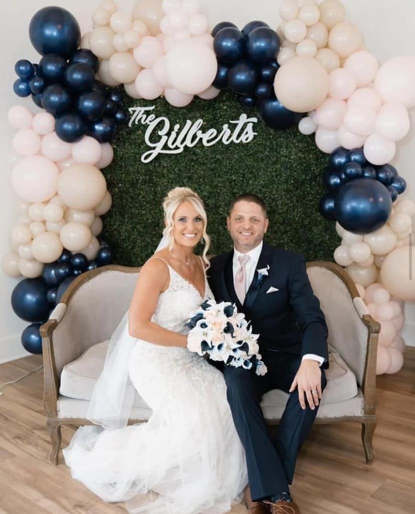 A bride and groom are sitting on a couch in front of a wall of balloons.