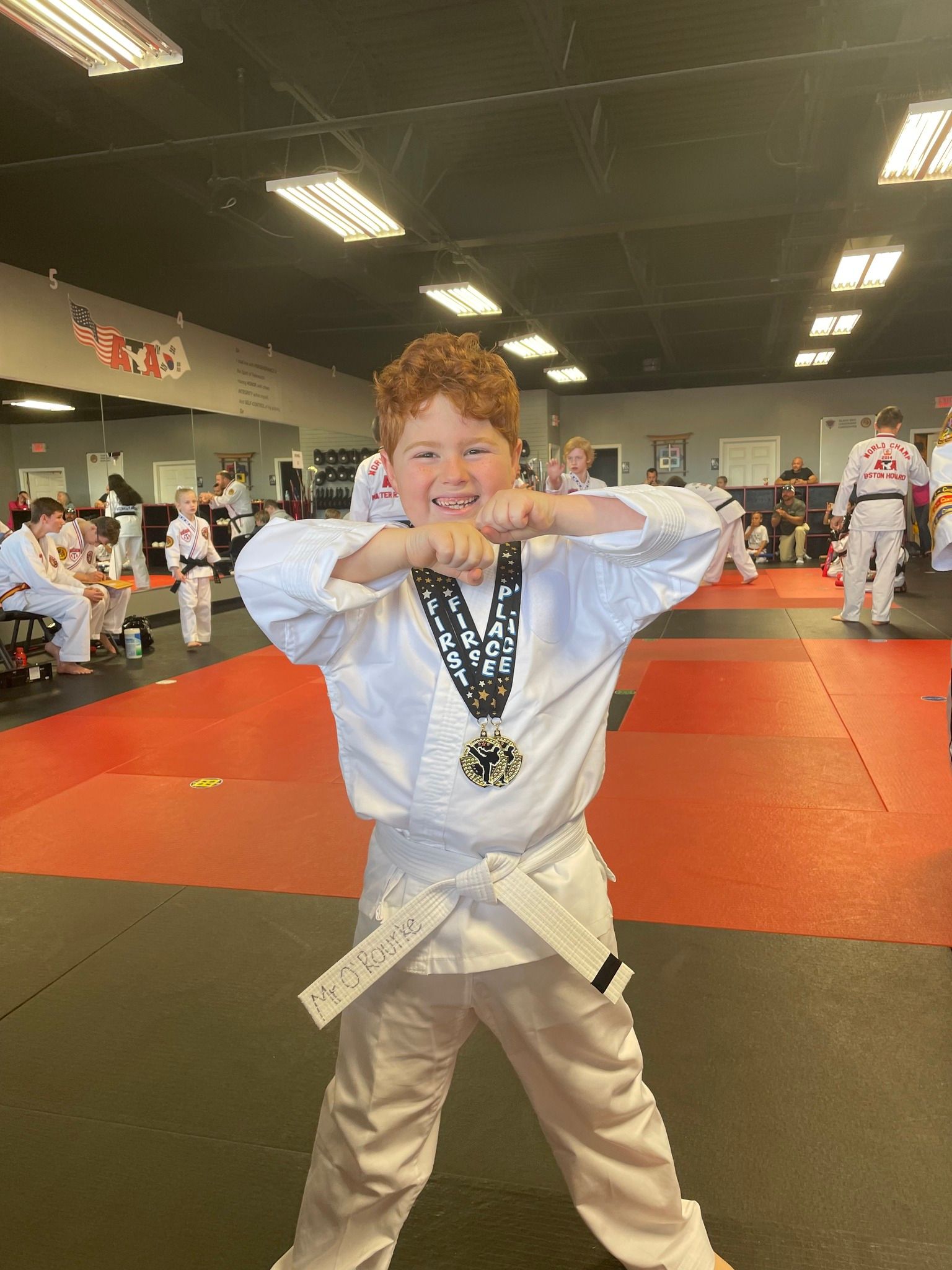 A young boy in a white karate uniform is standing in a gym with a medal around his neck.