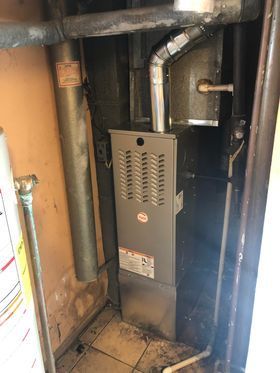 A silver furnace is installed in a small alcove with ductwork and pipes; beige walls.