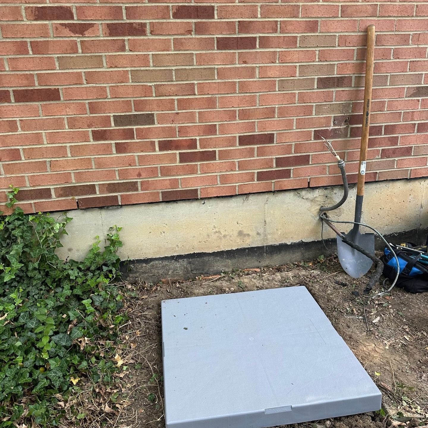 Grey rectangular cover on the ground near a brick wall. A shovel leans against the wall next to electrical wires.