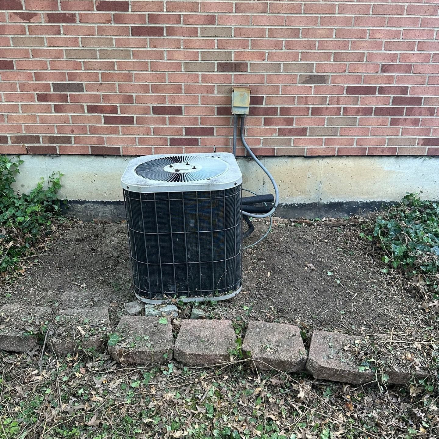 AC unit against a brick wall, with dirt and stone borders in front.