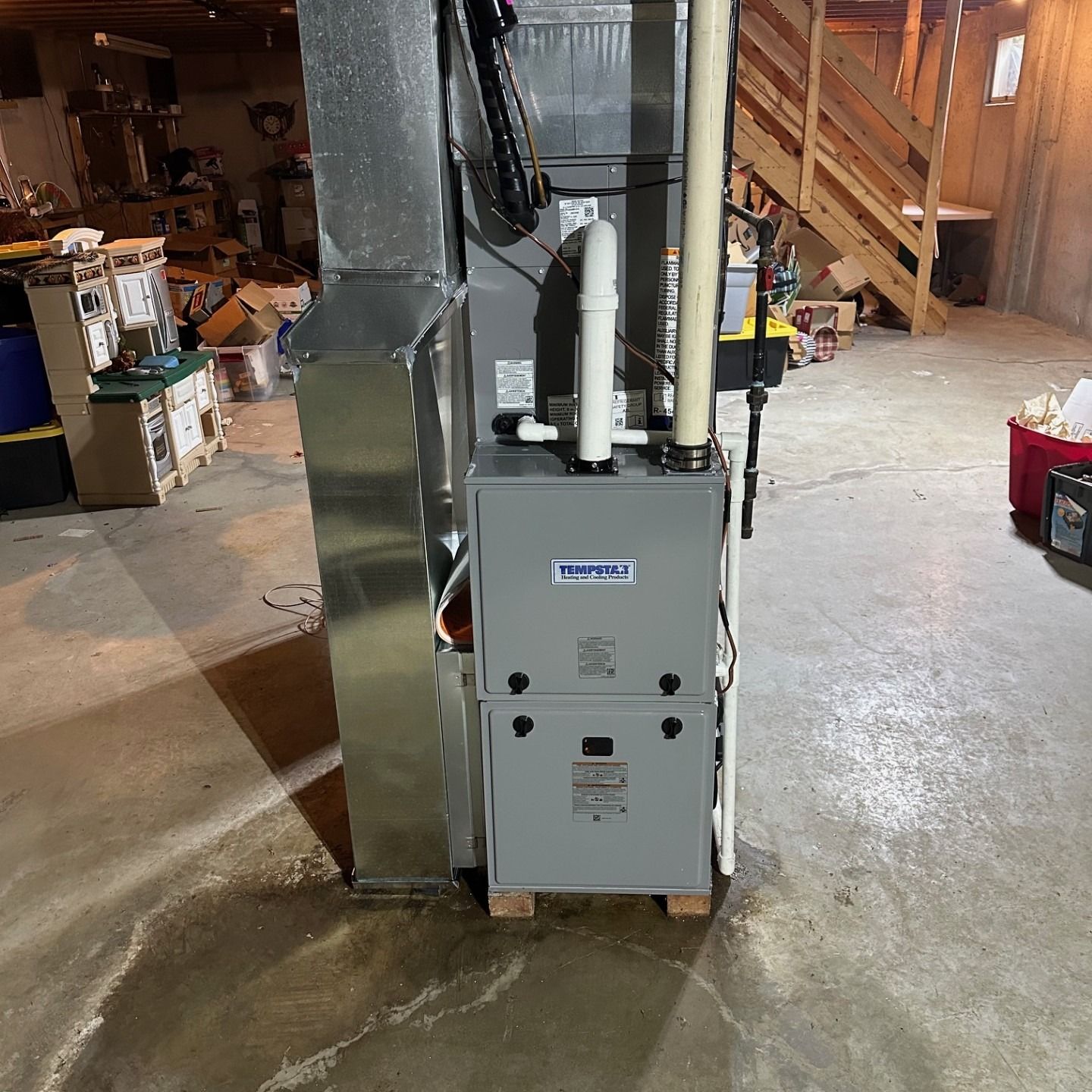 Furnace in a basement, with ductwork. Gray metal unit, pipes, wood stairs and concrete floor.