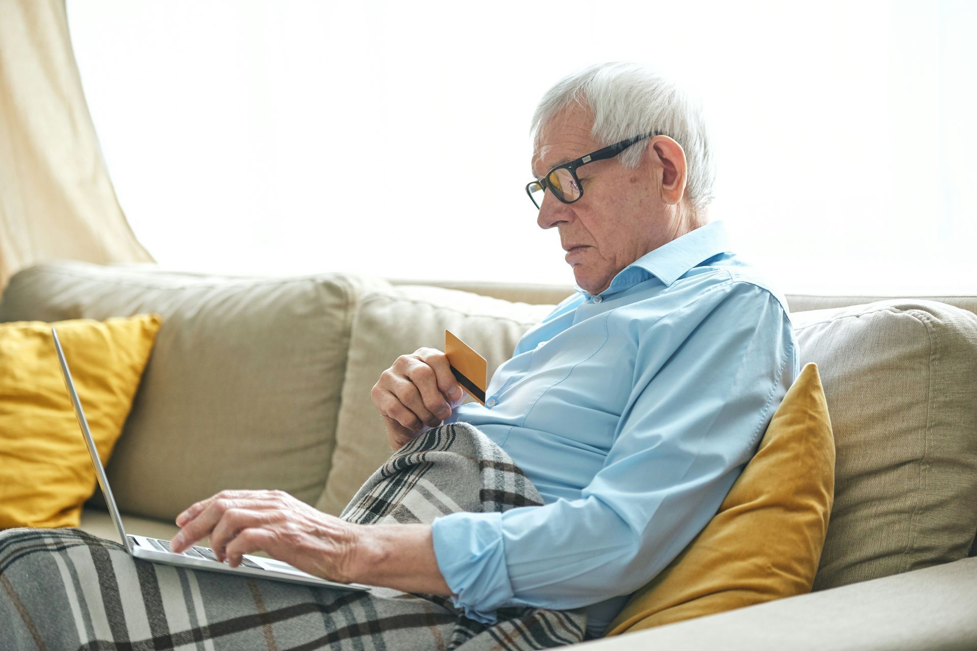 An elderly man is sitting on a couch using a laptop computer and holding a credit card.