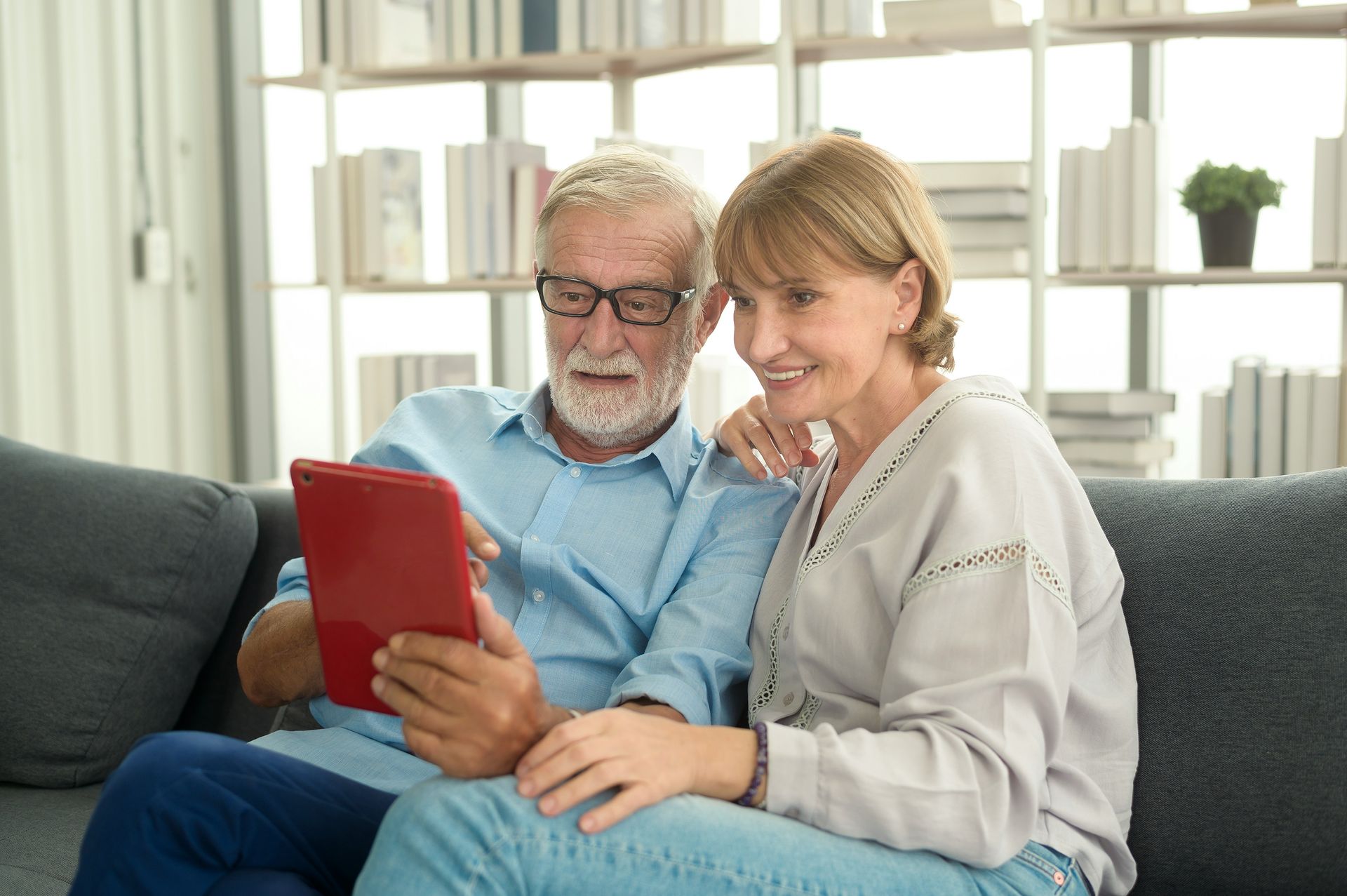 An elderly couple is sitting on a couch looking at a tablet.