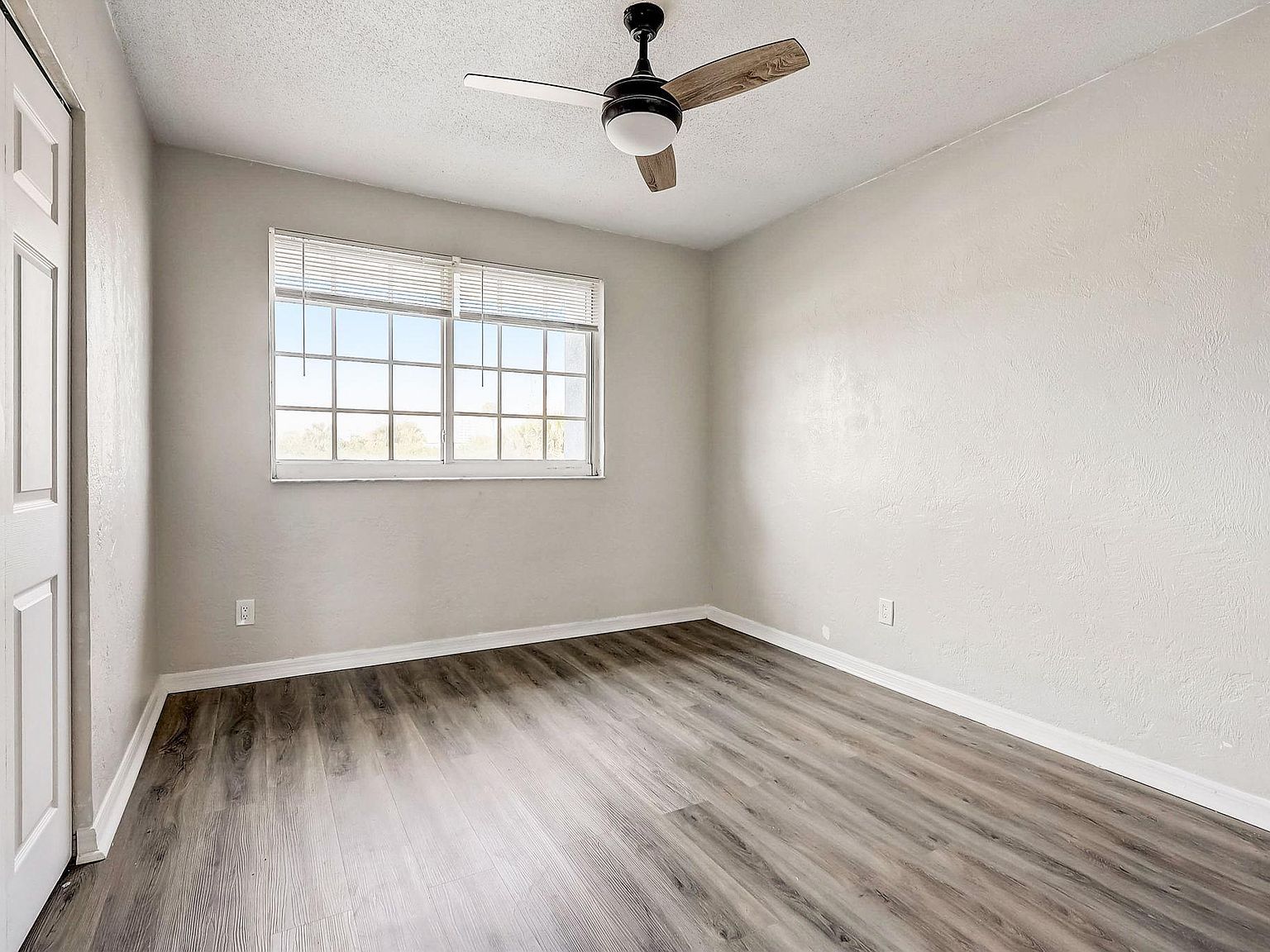 Empty room with gray-toned wood flooring, a white door, a window, and a ceiling fan.
