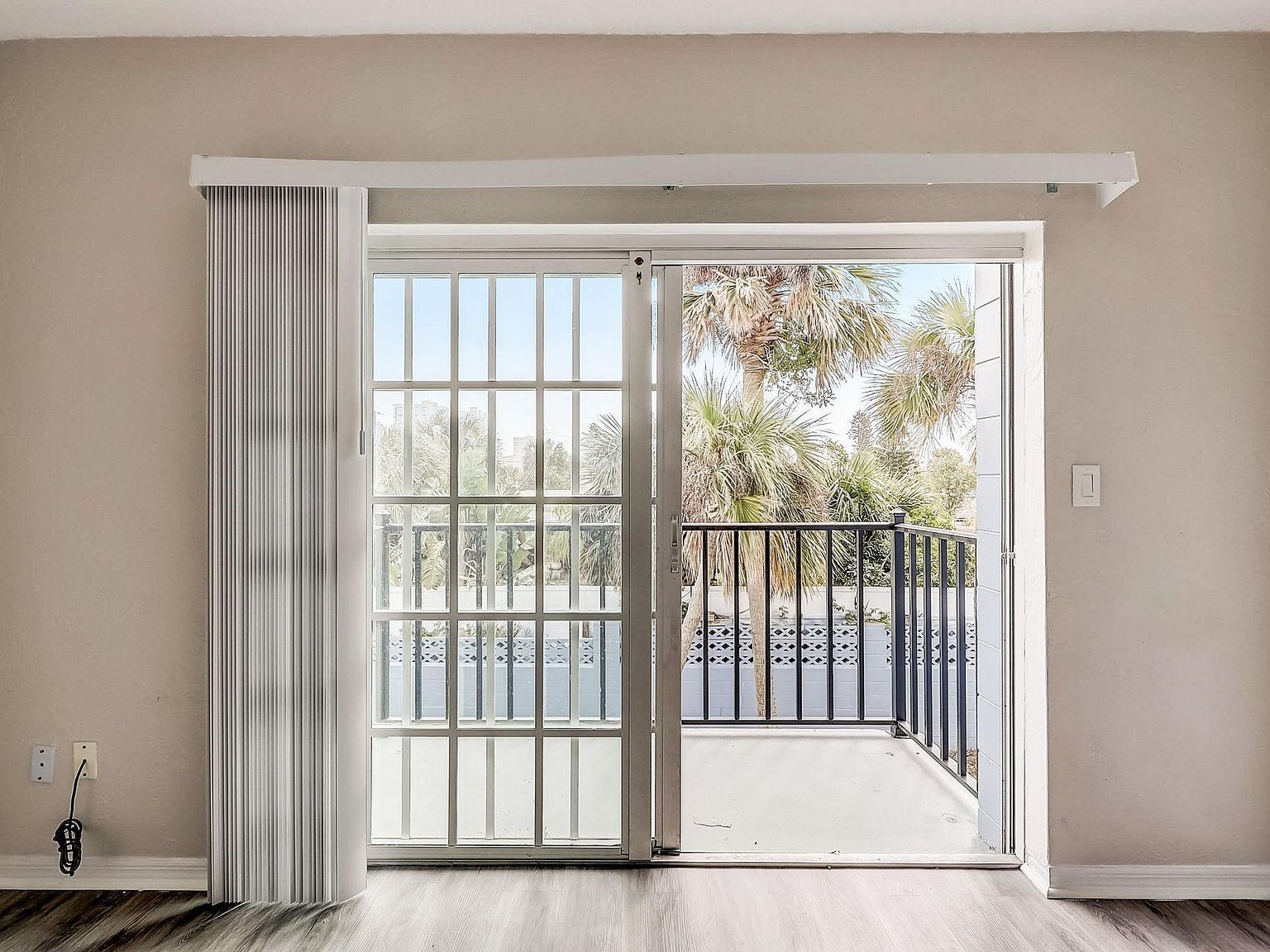 Sliding glass door leading to a balcony. White vertical blinds on the left. Trees visible outside.