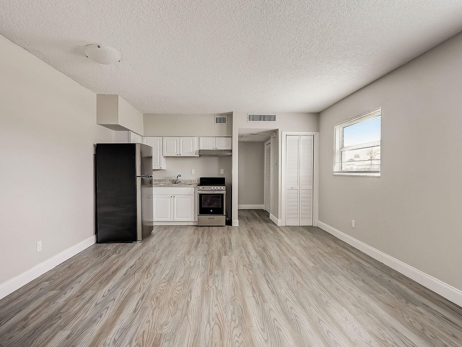 Empty apartment interior with kitchen and window. Gray walls, wood-look floor, black fridge.