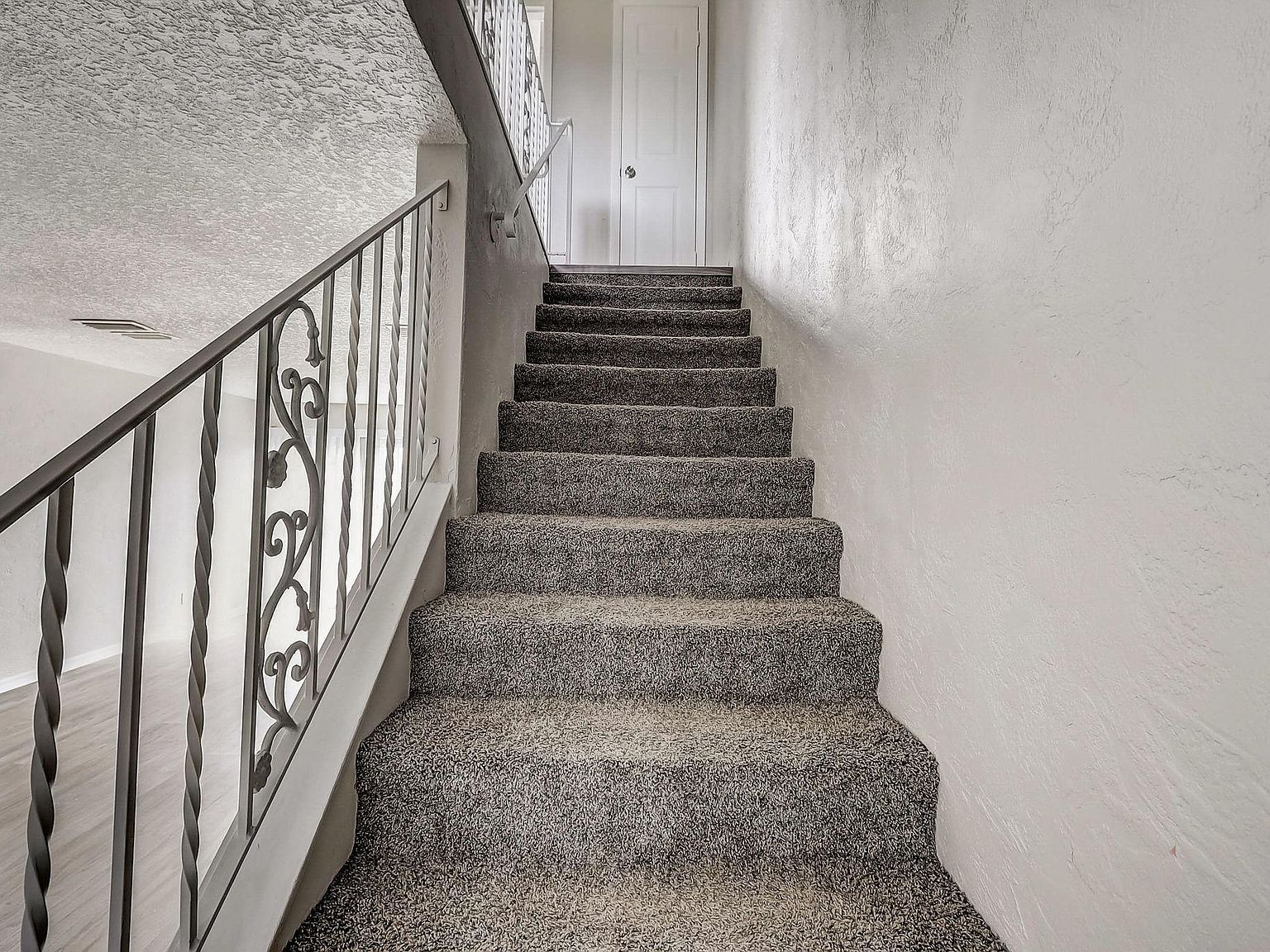 Staircase with gray speckled carpet, white walls, and a wrought-iron railing leading up to a closed white door.