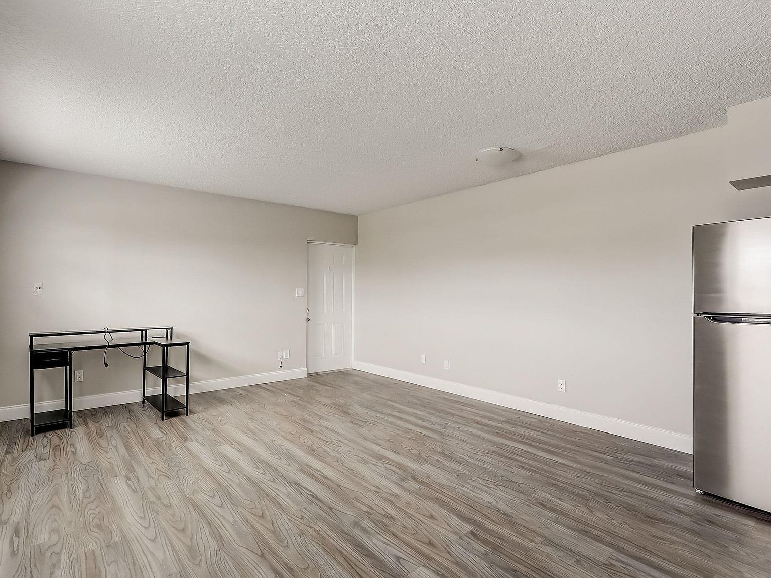 Empty, light-filled room with gray wood-look flooring, white walls, black desk, and stainless steel refrigerator.