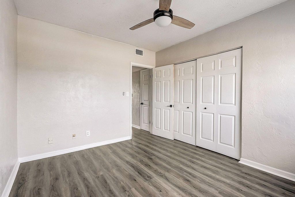 Empty bedroom with wood-look flooring, white walls and doors, a ceiling fan, and a glimpse of another room.