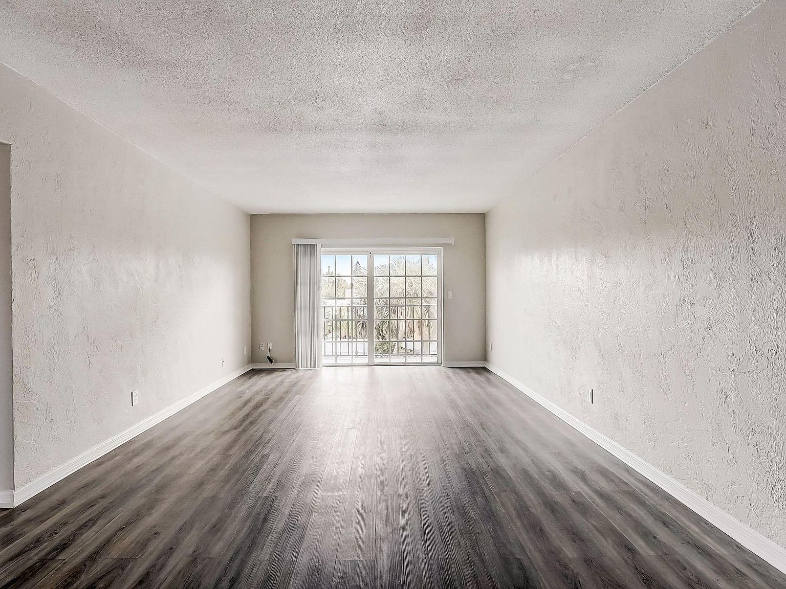 Empty living room with dark wood-look flooring, white walls, and a sliding glass door.