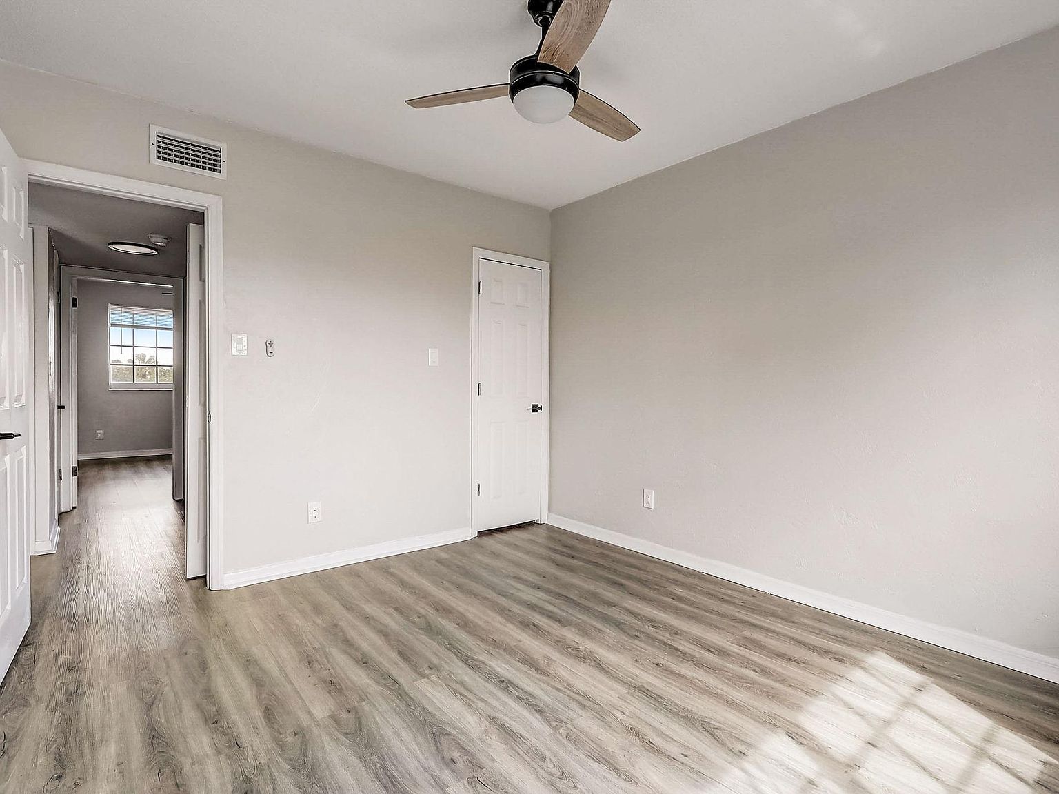 Empty room with gray walls, wood-look floor, ceiling fan, and a hallway.