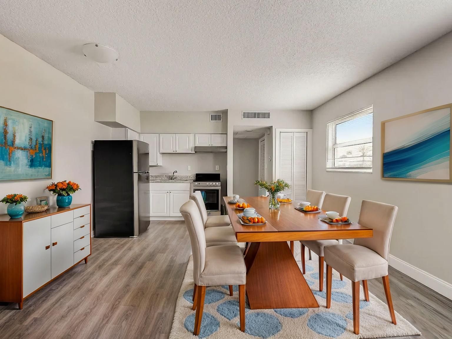 Dining room and kitchen. Dining table with eight chairs, blue and white rug, white cabinets, and artwork.