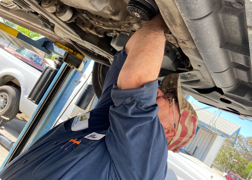 Mechanic under a car, on a lift, working. He wears a blue uniform and a camouflage hat.