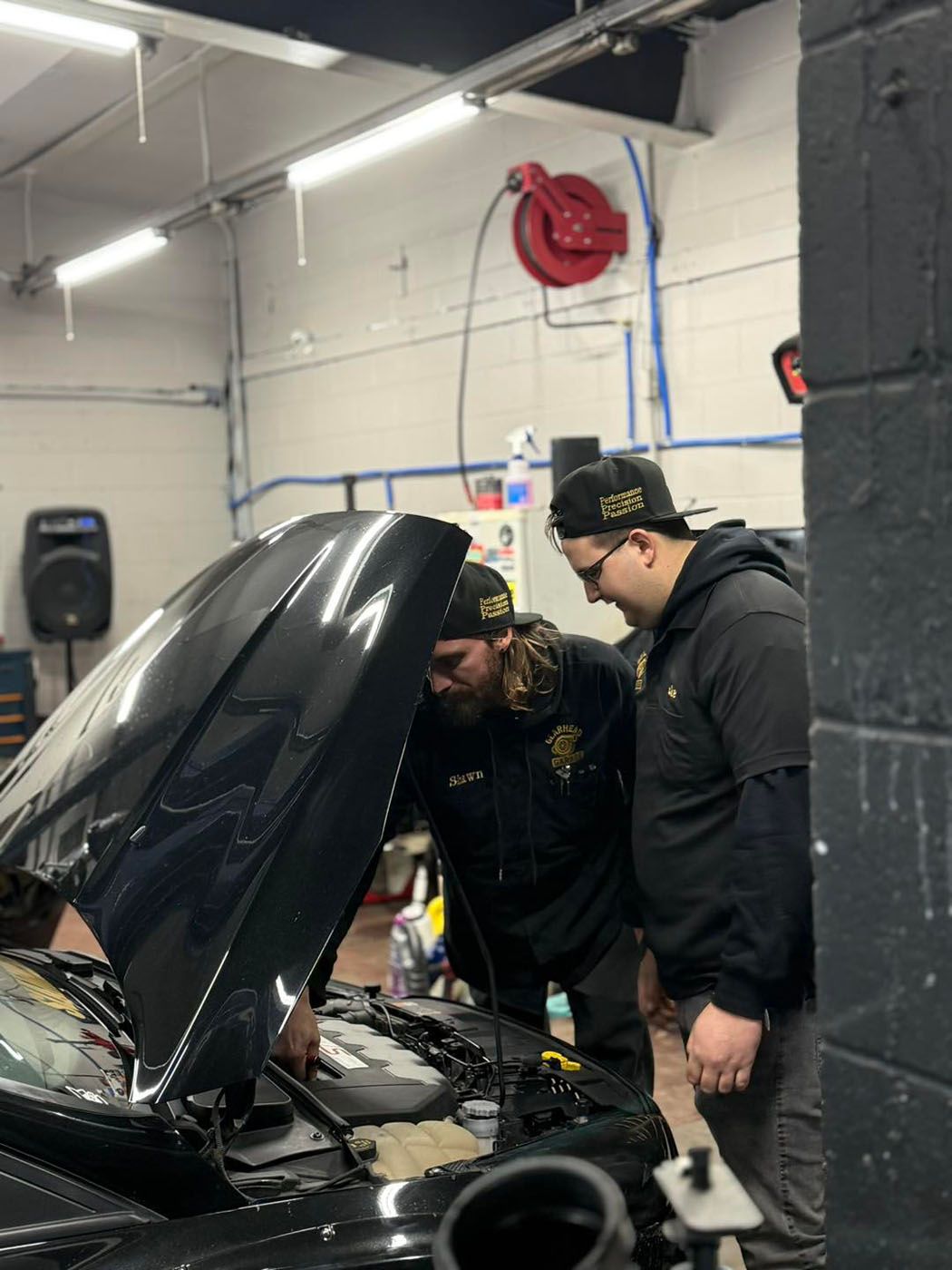 Two people in a garage lean over a car with its hood open, inspecting the engine.