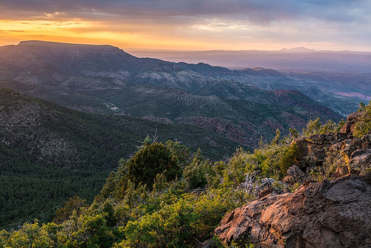 Sunset over forested mountains; golden sky, green trees, rocky foreground.