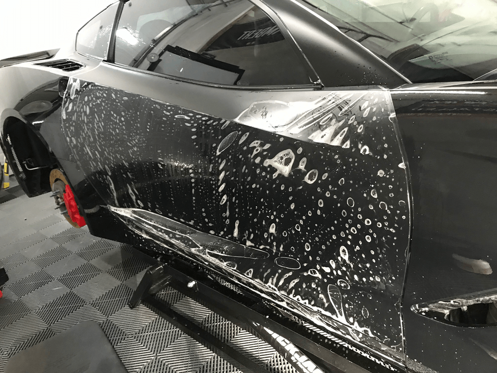 A black car is covered in a clear plastic film.